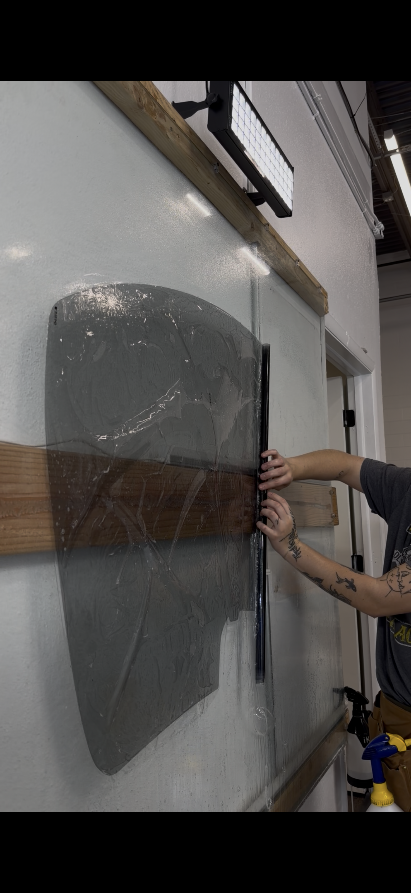 A person installs a gray, translucent window tint film onto a glass surface mounted over a horizontal wooden plank.