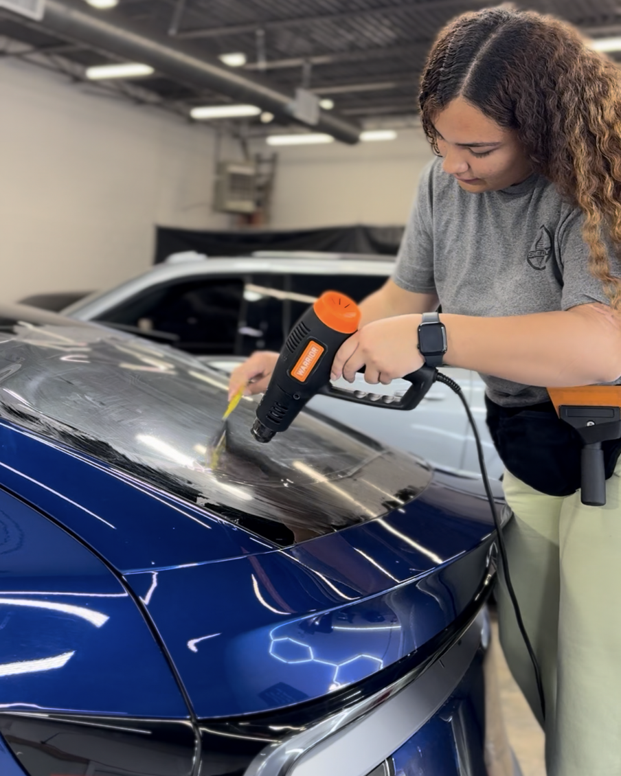A person using a heat gun to apply window tint to the rear windshield of a blue car in a garage.