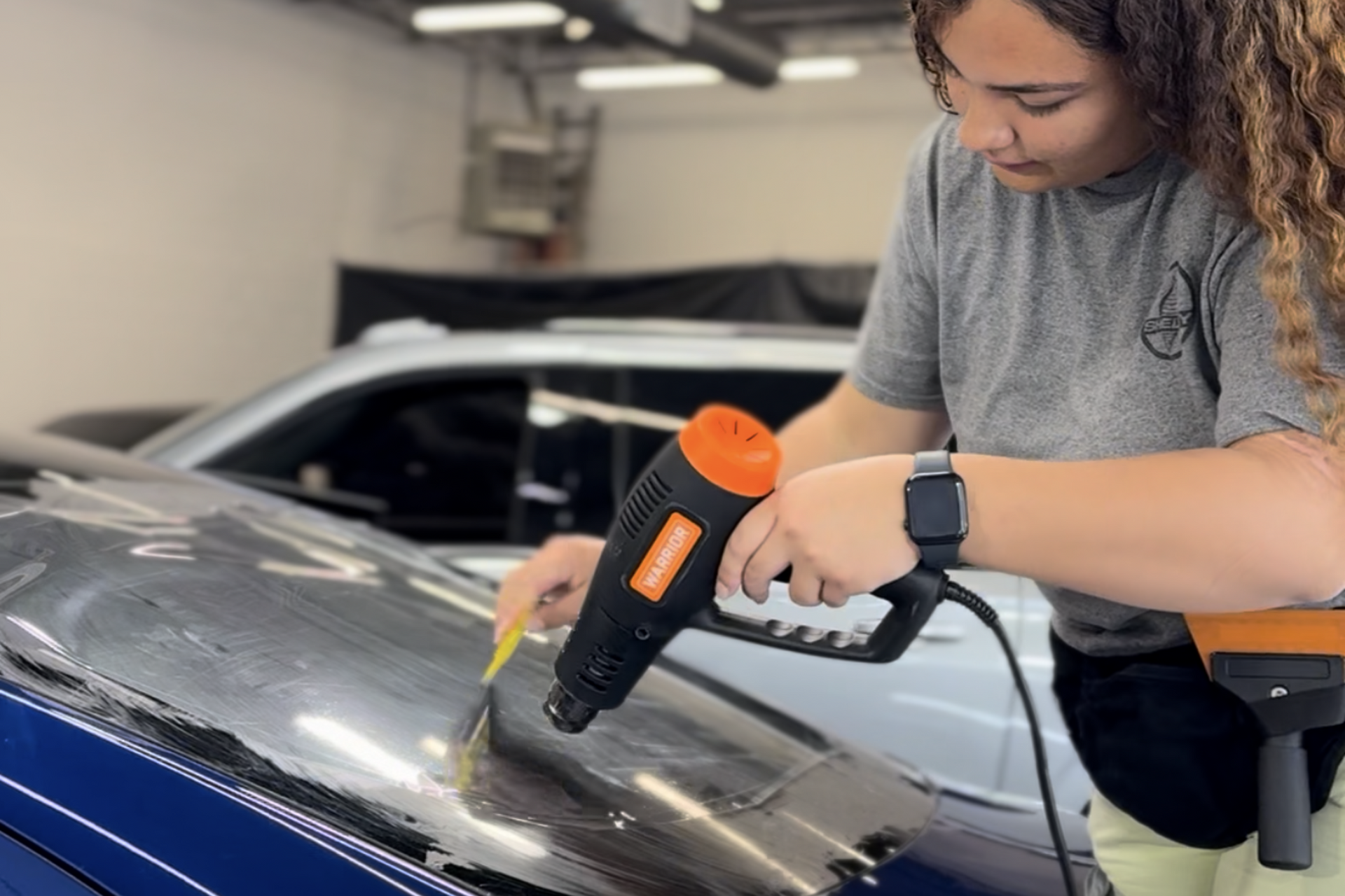 Technicians apply a clear protective film onto the hood of a black car in a garage with specialized overhead lighting.