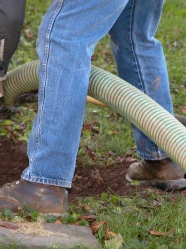Worker’s legs beside a large hose on muddy grass, wearing jeans and boots.