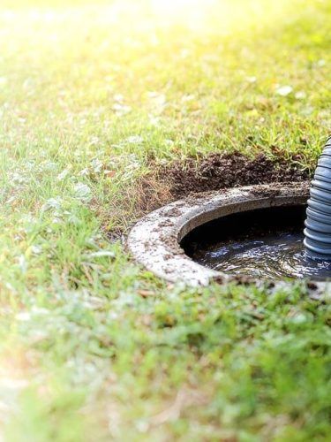 Open manhole in grassy ground with a black corrugated pipe nearby