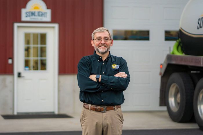 Smiling man in a dark shirt stands with arms crossed in front of a building and truck.