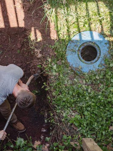 Person using a hose to water a garden bed beside a blue utility cover.
