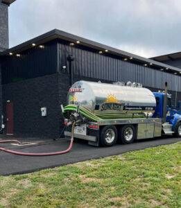 House with large white tanker truck parked on gravel driveway and lawn near woods