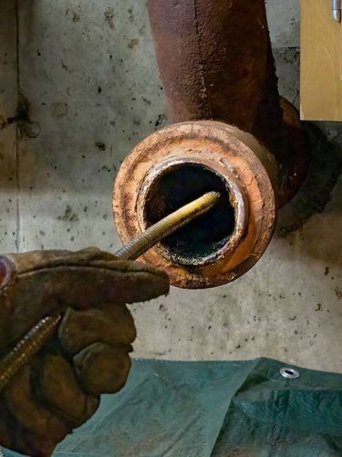 Gloved hand cleaning rust from a corroded metal pipe fitting with a tool