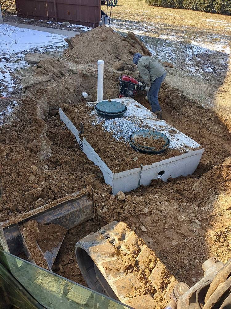 Man digging around a septic tank installation with white lids and PVC pipe in a dirt yard