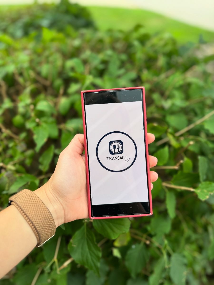 Hand holding a phone displaying a Transact app logo against a backdrop of green foliage.