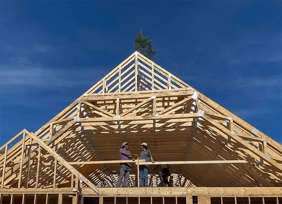 Two construction workers stand on the wooden frame of a roof under a clear blue sky.