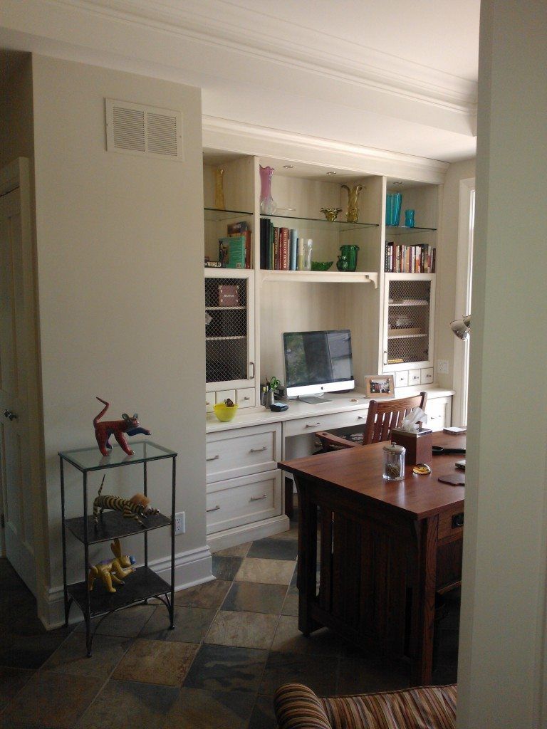 A home office with white built-in shelving, a dark wood desk, computer monitor, and a metal shelf with animal figurines.