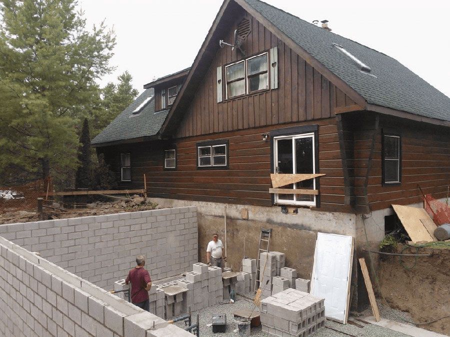 Two people construct a concrete block foundation wall next to a multi-story brown wooden house.