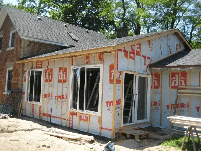 An addition under construction on the side of a brick house, covered in Tyvek wrap with new windows and a sliding door.