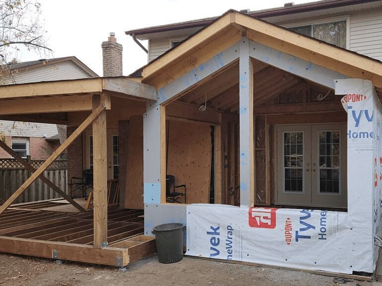 A house addition under construction, featuring a wooden deck frame and a framed structure covered in Tyvek house wrap.