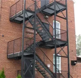 A tall, red brick building exterior featuring a black door with stairs, several windows, and a stone foundation.