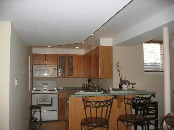 A compact basement kitchen with light wood cabinets, white appliances, a small peninsula with two stools, and a window.