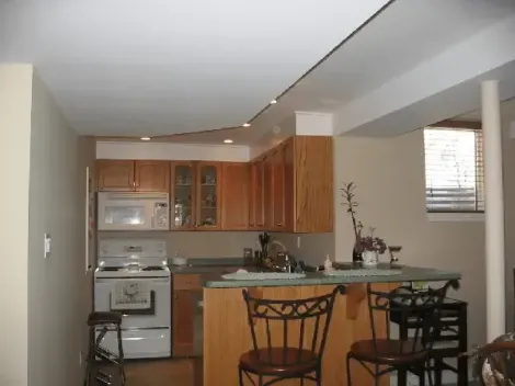A compact basement kitchen with light wood cabinets, white appliances, a small peninsula with two stools, and a window.