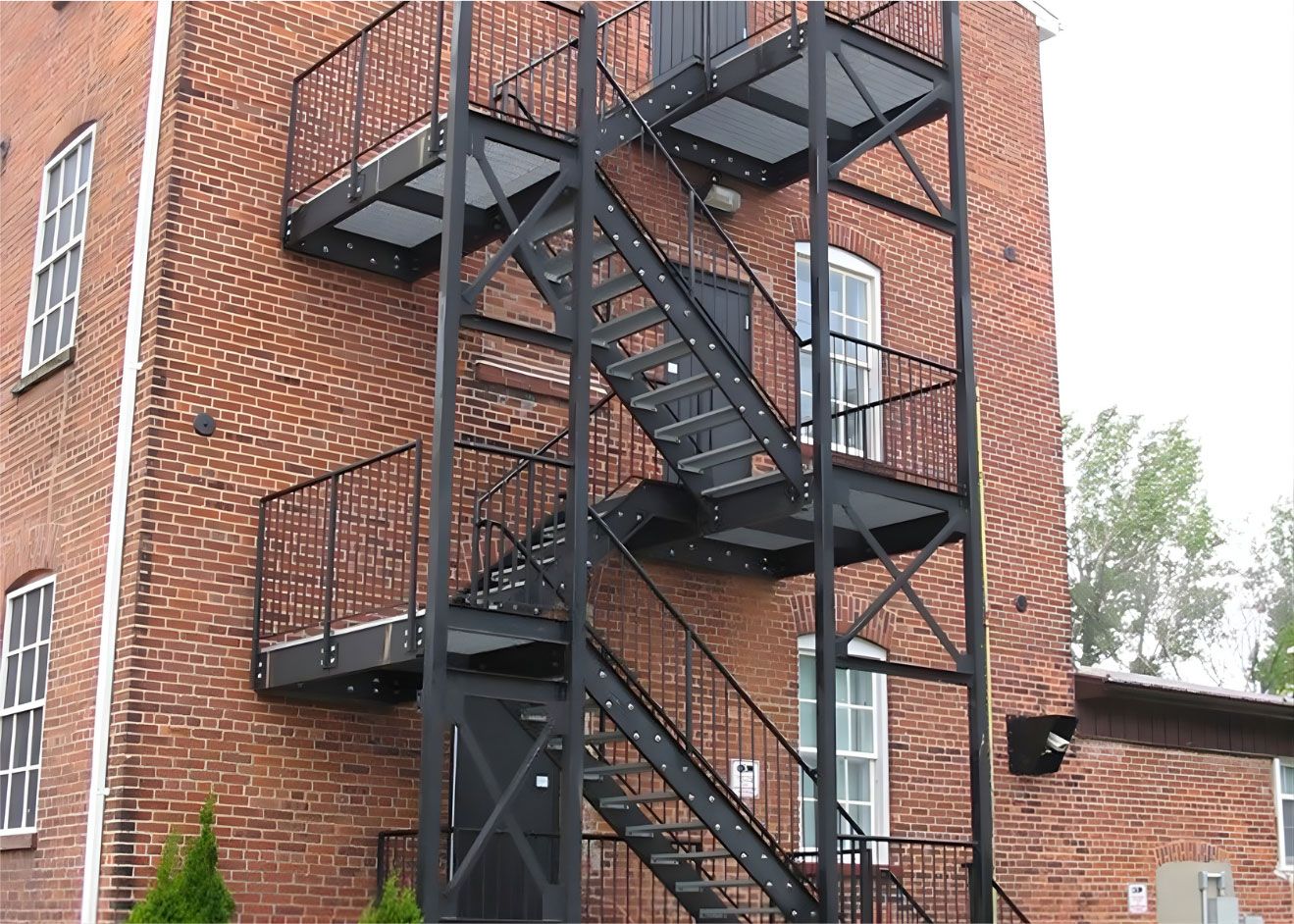 A tall, red brick building exterior featuring a black door with stairs, several windows, and a stone foundation.