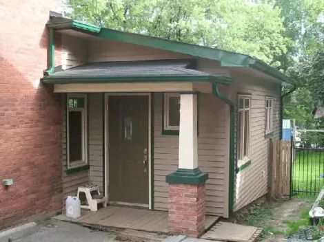 A small, beige, wood-sided building with a green-trimmed, sloped roof, an entrance door, and a brick pillar porch.