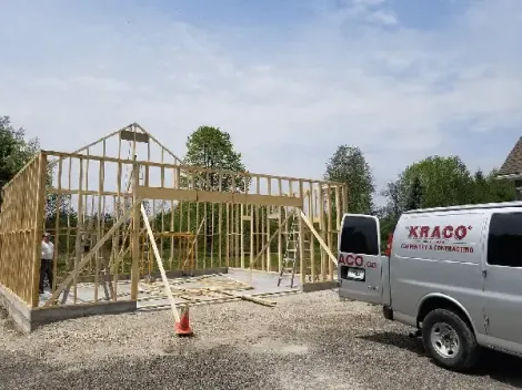 A wooden building frame under construction with a Kraco construction van parked in the foreground on a gravel lot.