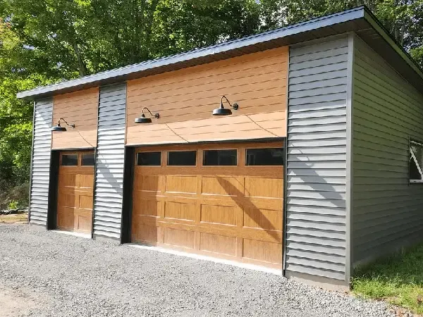 A modern detached two-car garage with gray horizontal siding and two wood-finish garage doors with overhead lamps.