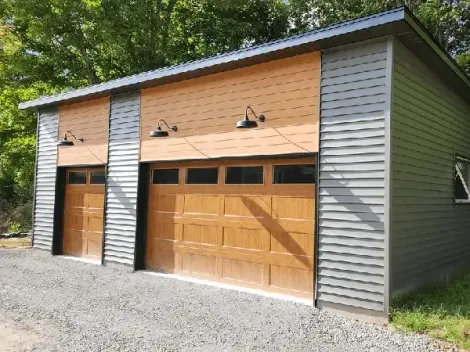 A modern detached two-car garage with gray horizontal siding and two wood-finish garage doors with overhead lamps.