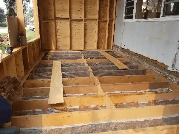 Construction site showing floor joists with insulation and plastic vapor barrier installed in an unfinished room.