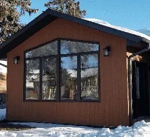 A brown, wood-paneled building with a gabled roof and large, multi-paned windows, surrounded by snow on a sunny day.