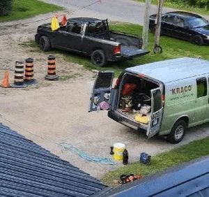 A high-angle view shows a work van with open doors, a black pickup truck, and traffic cones parked on a gravel lot.