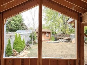 A view from inside a wooden building frame, looking out through large windows at a backyard with trees and a small shed.