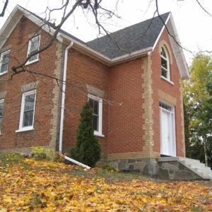 A two-story red brick building with white trim and a stone foundation, set among autumn leaves on a cloudy day.