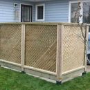 A light-wood privacy screen with diagonal lattice panels sits on a concrete base in front of a blue-sided house.