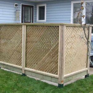 A light-wood privacy screen with diagonal lattice panels sits on a concrete base in front of a blue-sided house.