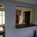 A wall with an open pass-through window looking into a room with furniture, a framed picture, and a doorway.