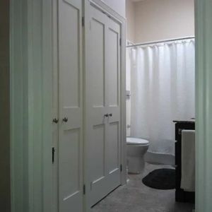 A view of a bathroom doorway featuring white double doors, with a toilet, shower curtain, and vanity visible inside.