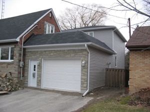 A two-story house addition featuring a stone-front garage, white door, and grey siding, attached to a brick home.