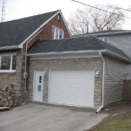 A garage with light-colored stone facing, a white door and garage door, and gray siding, attached to a brick house.