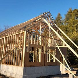 A wooden house frame under construction on a concrete foundation, featuring roof trusses against a clear blue sky.