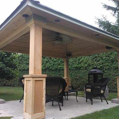 A wooden gazebo over a stone patio with wicker chairs and a fire pit in a grassy yard.