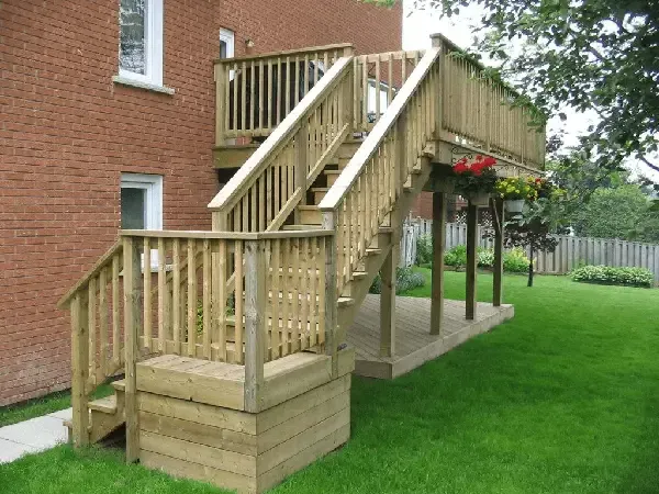 Wooden deck attached to a brick house with stairs leading down to a lower patio area and a grassy backyard.