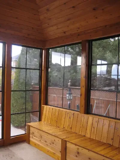 A sunroom with wooden walls, a ceiling, and bench seating, featuring large, multi-pane windows overlooking a yard.