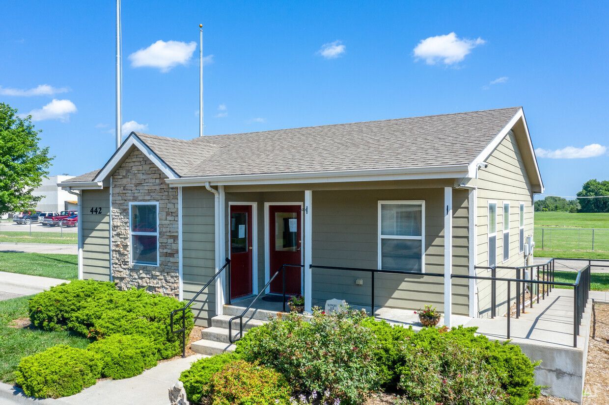 Small, beige building with stone accent, red doors, and ramp for accessibility. Lush bushes in front.