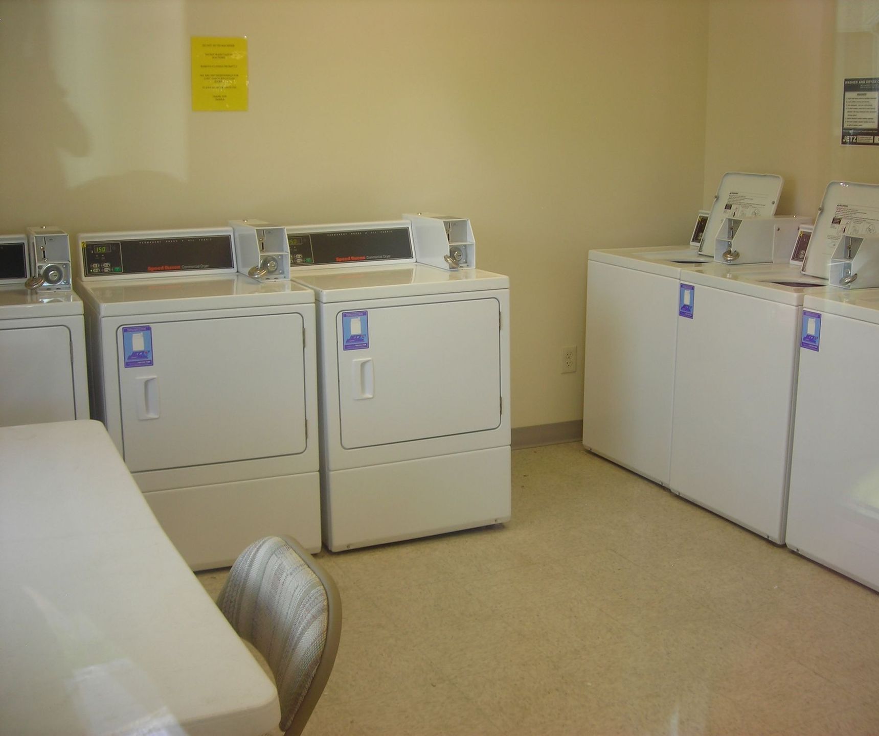 Laundry room with white washing machines and dryers; table and chair in foreground.