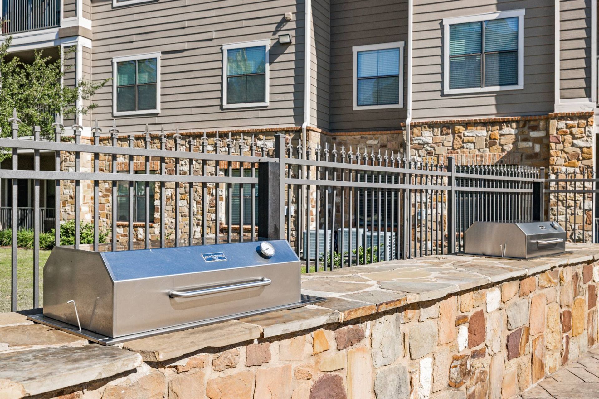 Outdoor communal grilling area with stainless steel grills along a stone ledge beside an apartment building.