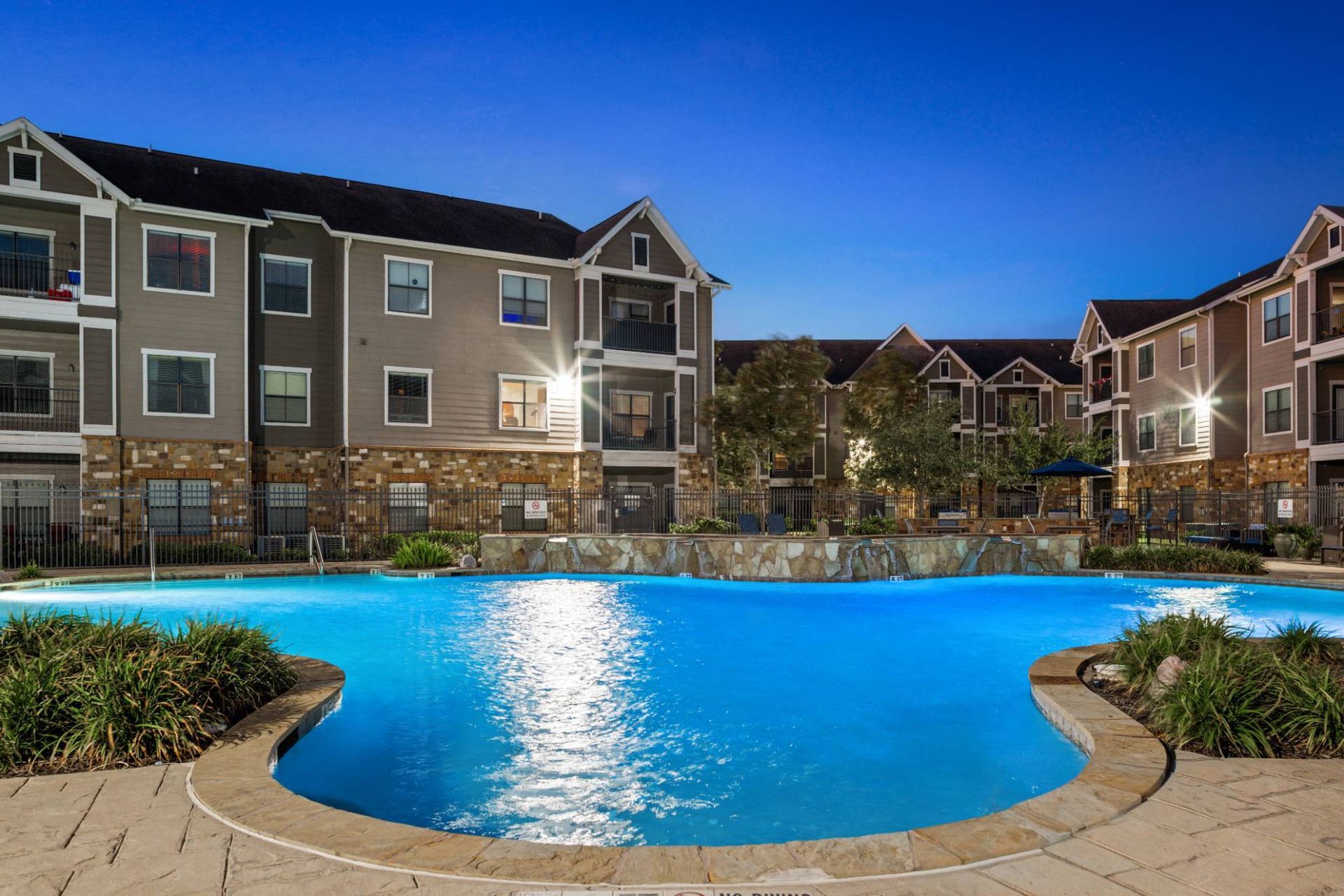 Evening view of an outdoor apartment community pool with surrounding buildings and lounge chairs.