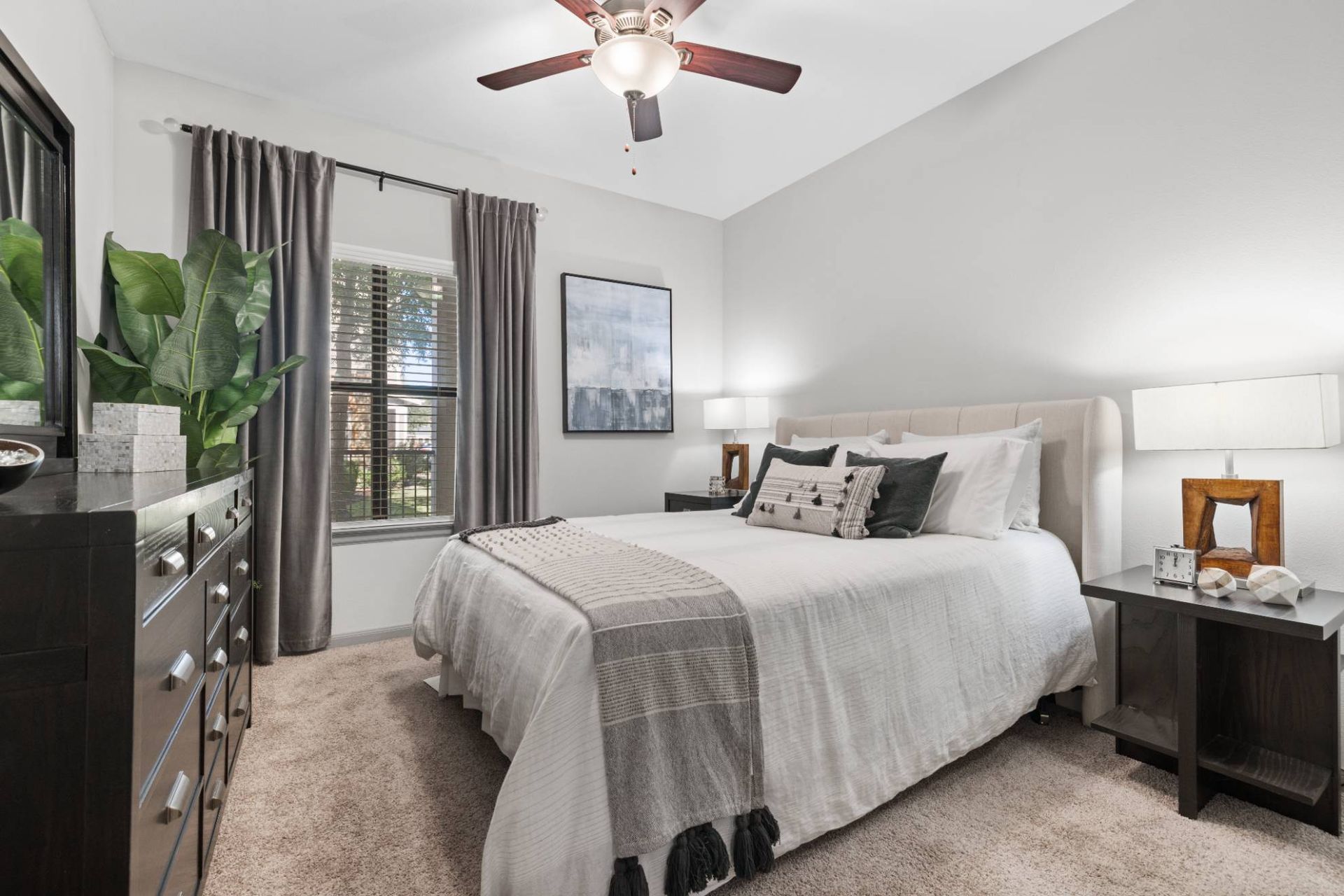 Bedroom in an apartment with a large bed, two nightstands, a dresser, a plant, and a window with curtains.