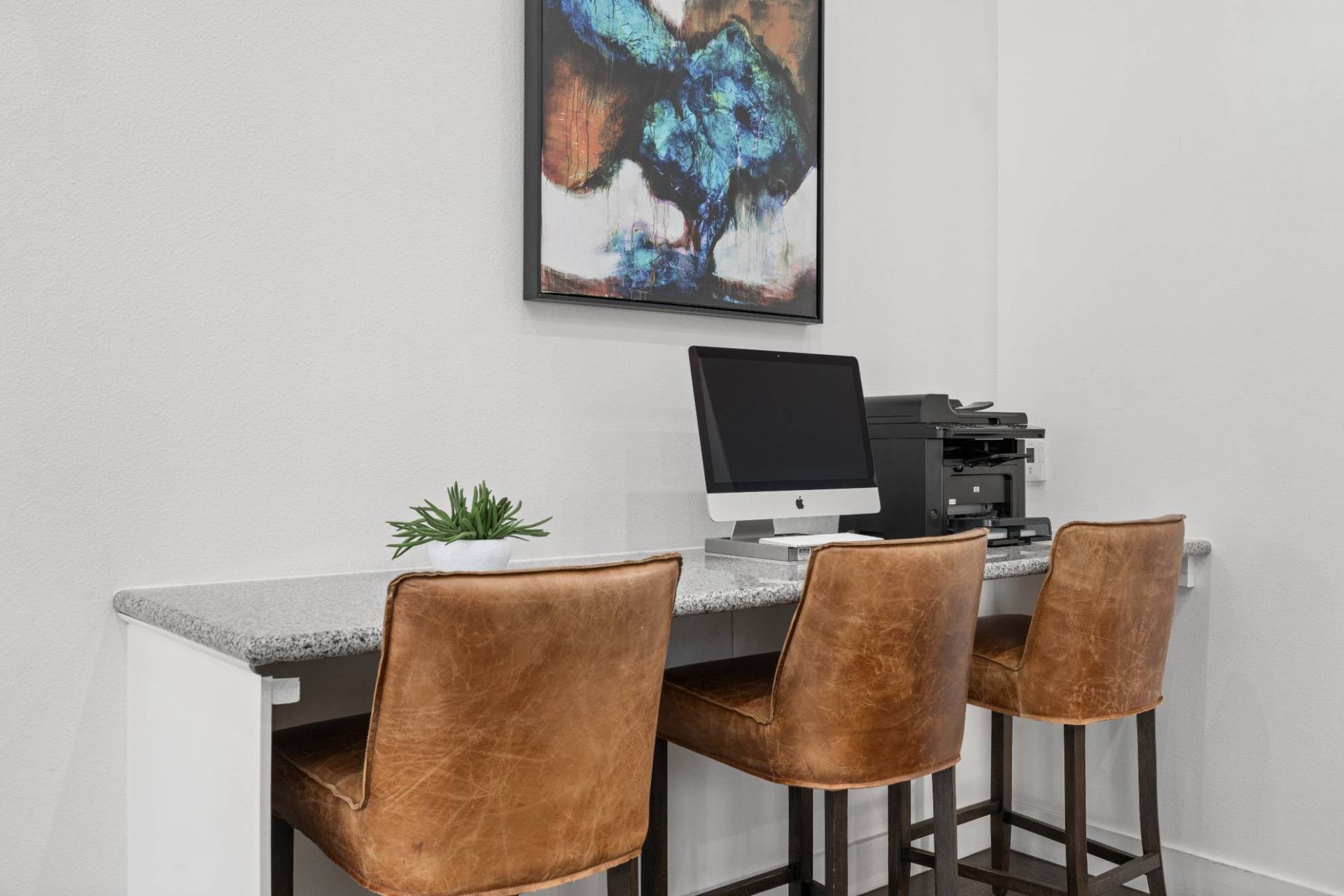 Leasing-office workspace with a granite counter, iMac, printer, plant, and three brown leather stools.