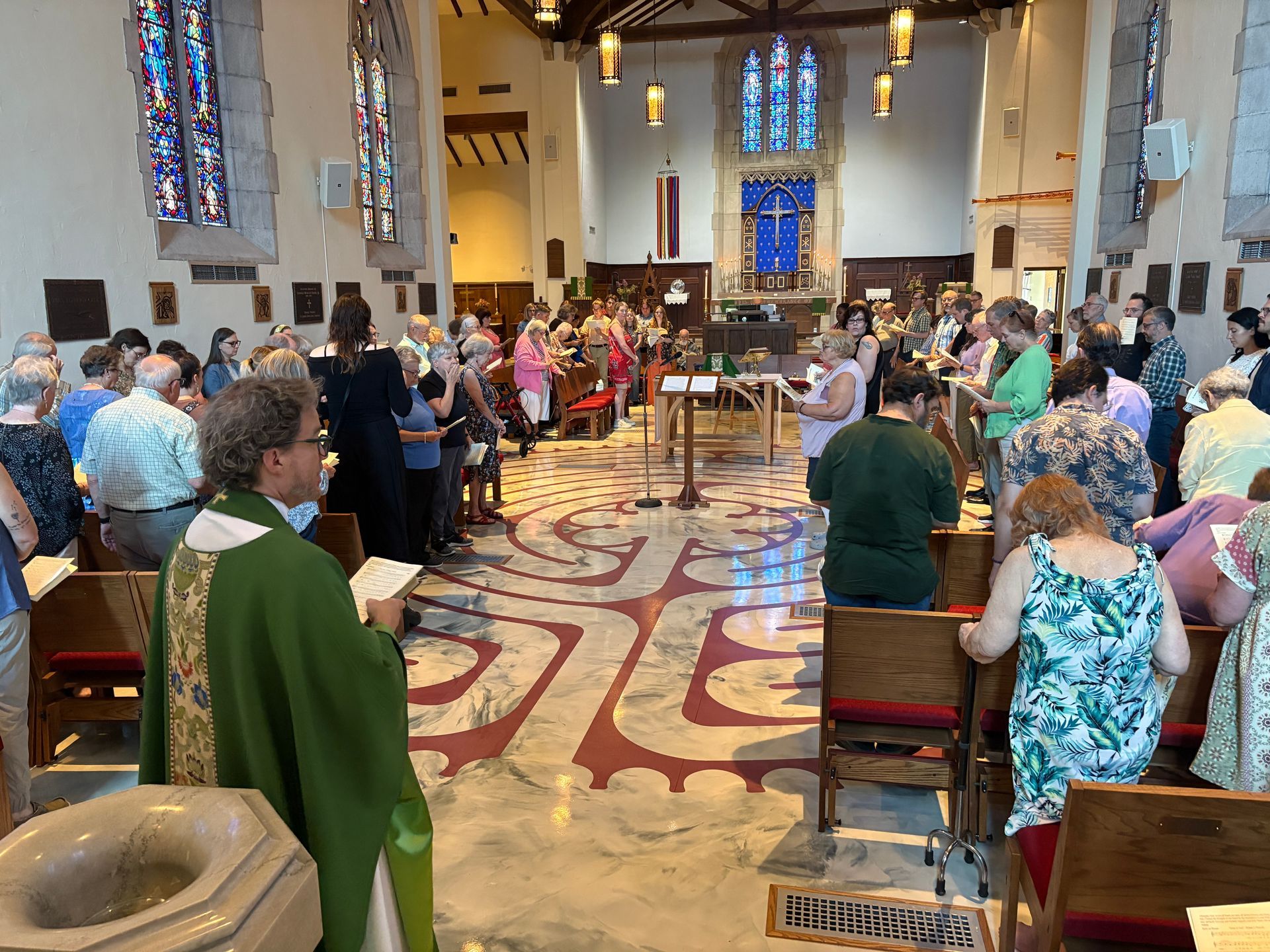 Church service with people standing, seated, or kneeling. Priest in green vestments. Stained glass windows and decorative floor.