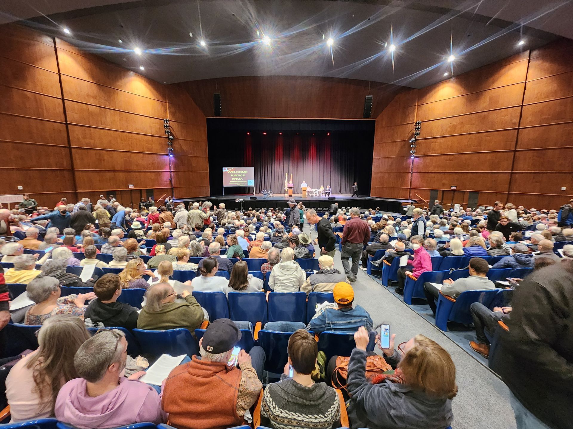 Large audience in a theater, facing a stage with people. Many seated, some taking photos.