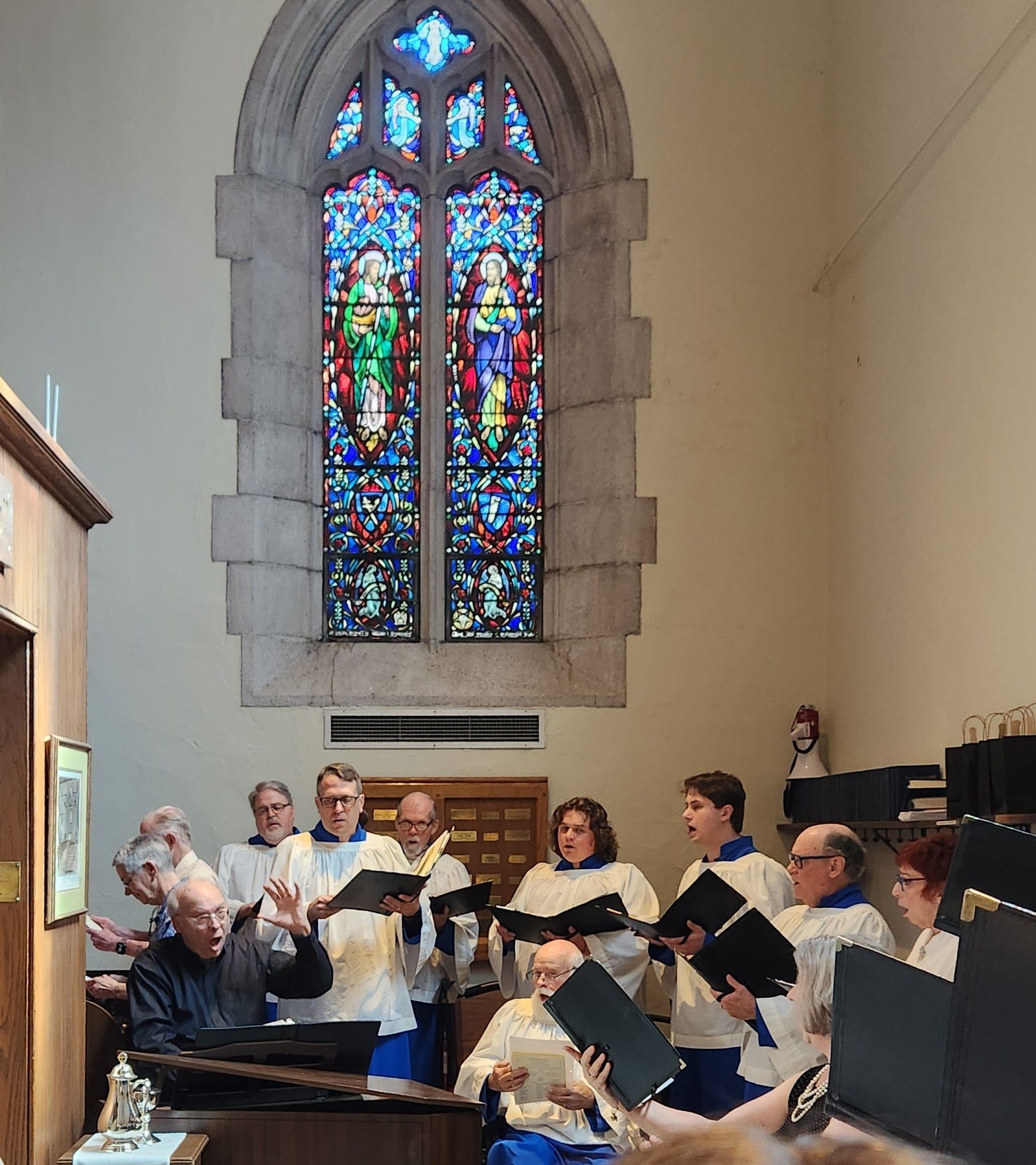 Choir singing in front of a stained-glass window. People in white robes and blue collars, holding music.