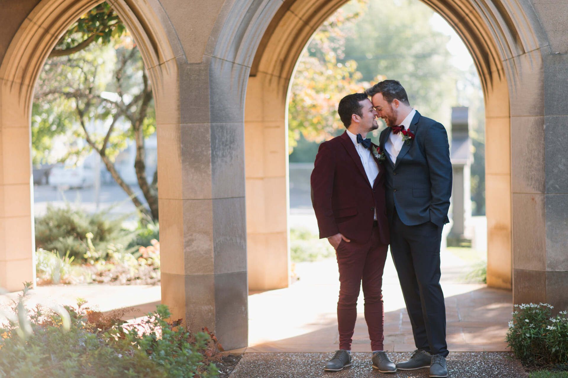 Two men in suits share a kiss under stone arches, bathed in sunlight.