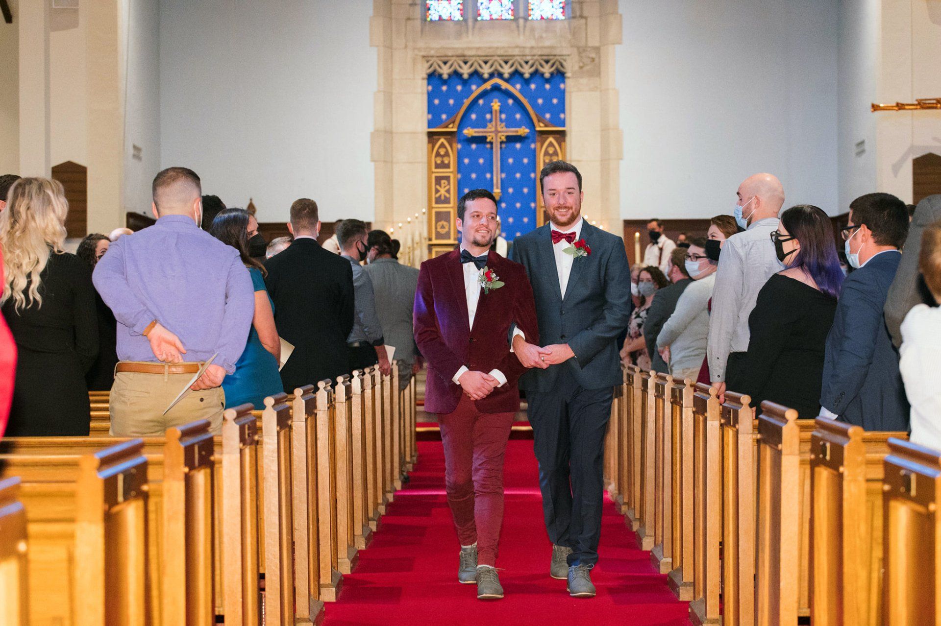 Two grooms in suits walk down the aisle of a church during a wedding ceremony.